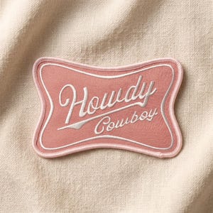 May include: A pink embroidered patch with the words "Howdy Cowboy" in white script. The patch has a curved rectangular shape with a white border and a pink outline. The patch is on a cream-colored fabric background.