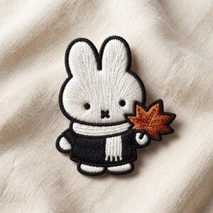 May include: An embroidered patch featuring a white rabbit character with black eyes and a cross-shaped mouth. The rabbit wears a black sweater and a white scarf, and holds a single orange-brown maple leaf in one hand, outlined in black thread.
