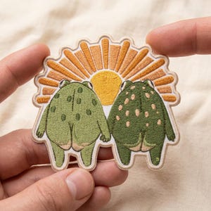May include: An embroidered patch featuring two green frogs standing side-by-side, holding hands, and facing away from the viewer. Above the frogs is a yellow and orange sunburst design. The patch has a light beige border and a scalloped top edge.