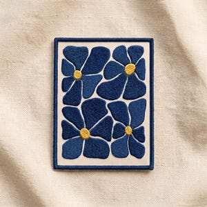 May include: A rectangular embroidered patch featuring a cream-colored background with a pattern of four stylized dark blue flowers. Each flower has rounded petals and a small, circular, golden-yellow center. The patch has a dark blue stitched border.