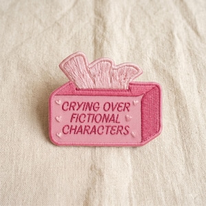 May include: A pink embroidered patch shaped like a tissue box with the text "Crying Over Fictional Characters." The patch features a stack of tissues and small heart details.