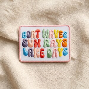 May include: Embroidered patch with the words "Boat Waves Sun Rays Lake Days" in colorful, bubble-style letters. The patch has a white background with a pink border, and is set against a neutral fabric.