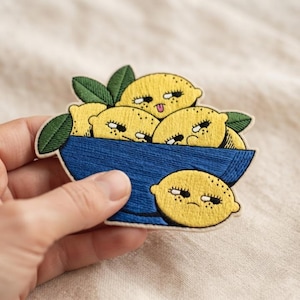 May include: An embroidered patch featuring a deep blue bowl filled with yellow lemons that have expressive faces, including one sticking its tongue out and others with skeptical expressions, accented by green leaves and a single lemon below.