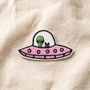 May include: Embroidered patch featuring a pink UFO with a green alien and a black cat inside. The UFO has a white window and is outlined in black. The patch is on a beige fabric background.