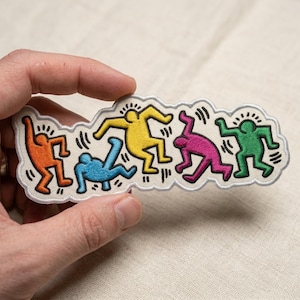 May include: An embroidered patch featuring five stylized human figures in motion, each in a different color: orange, blue, yellow, magenta, and green. The figures are outlined in black thread against a white background with a scalloped border.