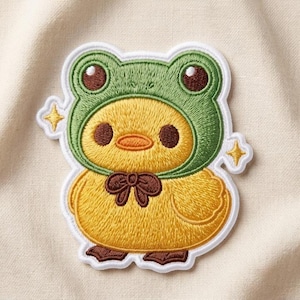 May include: An embroidered patch featuring a yellow duckling wearing a green frog hood. The duckling has dark brown eyes, an orange beak, and a brown bow tie. Two small yellow four-pointed stars are positioned on either side of the character.