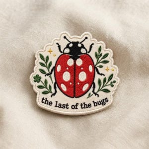May include: An embroidered patch featuring a red ladybug with white spots, black head, and legs, surrounded by green leaves and small yellow stars. The text "the last of the bugs" is stitched in black at the bottom on a cream-colored background.