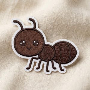 May include: An embroidered iron-on patch shaped like a brown ant with a large head, two antennae, and six legs. The ant features a smiling face with two large, round, dark eyes containing white highlights, all outlined in a thick white border.