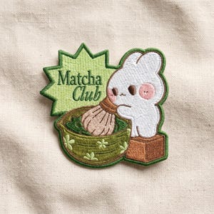 May include: Embroidered patch featuring a white bunny with pink cheeks, whisking matcha in a green bowl. The patch includes the text "Matcha Club" on a green starburst background. The patch is on a beige fabric.