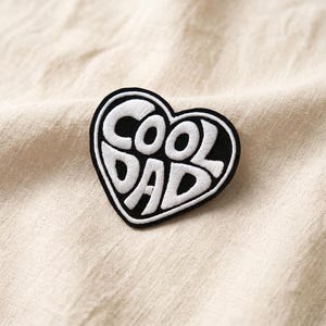 May include: A heart-shaped patch with the words "COOL DAD" embroidered in white on a black background. The patch is set against a beige fabric background, creating a simple yet striking design.
