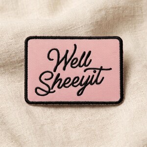 May include: A rectangular pink patch with the phrase "Well Sheeeyit" embroidered in black cursive script. The patch has a black border and is set against a neutral fabric background.