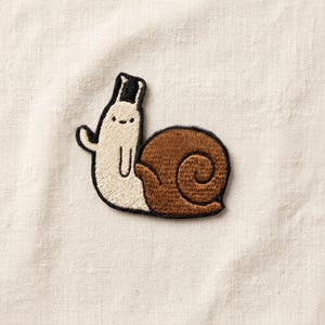 May include: Embroidered patch featuring a whimsical snail character. The snail has a cream-colored body, a brown shell, and is wearing a small black top hat. The patch is outlined in black and is designed to be ironed or sewn onto fabric.
