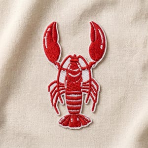 May include: A red lobster embroidered patch featuring white highlights on the claws and body. The design is rendered in dense red thread with a white border outline, showcasing the segmented tail, multiple legs, and two large raised claws.
