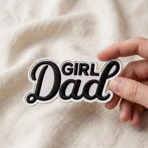 May include: Embroidered patch with the words "Girl Dad" in a black script font with a white border. The patch is held against a neutral fabric background.