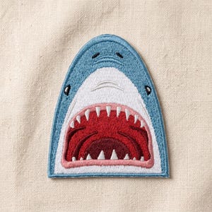 May include: An embroidered iron-on patch shaped like a shark head. The design features a light blue top, a white snout, and a wide-open mouth with sharp white teeth and a dark red interior. The patch has a stitched blue border and a pink gum line.