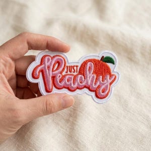 May include: An embroidered patch with a white border featuring the text "JUST Peachy" in a stylized pink and red script font. The design includes an orange peach graphic with a small green leaf, all set against a white fabric background.