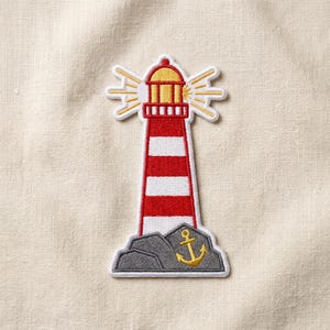 May include: An embroidered patch featuring a lighthouse with horizontal red and white stripes, a yellow lantern room, and radiating yellow light beams. The base consists of gray rocks with a small, yellow embroidered anchor icon on the front.