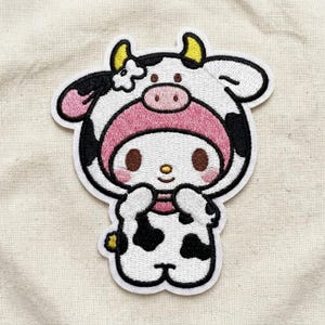 May include: An embroidered patch featuring a cartoon character wearing a cow costume. The character has a white face with brown eyes, a pink hood, and yellow horns. The cow suit is white with black spots, a pink snout, and a small white flower detail.