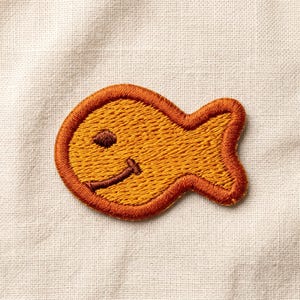 May include: Embroidered patch in the shape of a smiling fish. The patch is primarily orange with a brown outline, eye, and mouth. The patch is on a cream-colored fabric background.