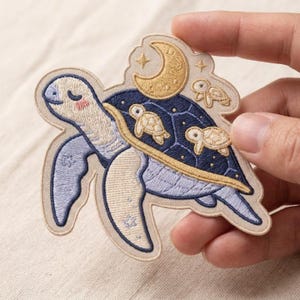 May include: An embroidered patch featuring a light blue sea turtle with a dark blue shell. The shell is decorated with two small cream-colored baby turtles and a golden crescent moon. The design includes small star accents and a cream border.