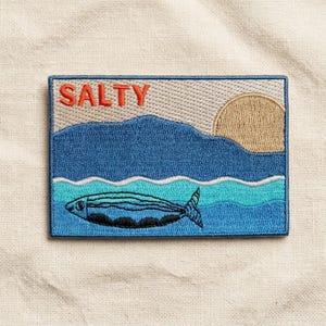 May include: A rectangular embroidered patch featuring the word SALTY in orange letters. The design depicts a blue mountain range, a tan sun, and a dark blue fish swimming in turquoise and white waves against a light beige background.