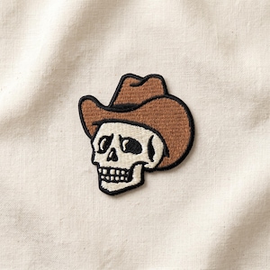 May include: Embroidered patch featuring a skull wearing a brown cowboy hat. The skull is white with black eye sockets and a jawline. The hat is brown with a black outline. The patch is on a cream-colored fabric.