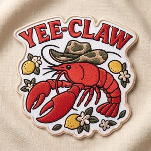 May include: An embroidered patch featuring a bright red crawfish wearing a tan and brown patterned cowboy hat. The text YEE-CLAW is arched above in red letters. The design includes yellow lemons and small white flowers on a white background.