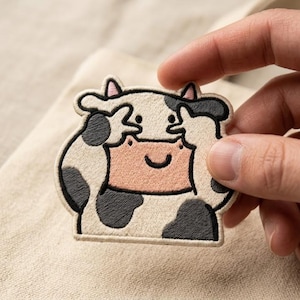 May include: An embroidered patch featuring a cartoon cow with black and white spots, pink inner ears, and a light pink muzzle. The cow has its hooves raised to its face, covering its eyes in a peek-a-boo gesture, with a simple black smile.
