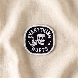 May include: A black and white patch with the words "EVERYTHING HURTS" surrounding a skull and crossbones design. The patch also features a thumbs-up hand gesture and lightning bolts.