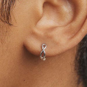 May include: A small silver hoop earring featuring an infinity symbol design. The earring is worn in the earlobe and has a polished metallic finish. The infinity loop is centered on the hoop, creating a continuous, sleek, and simple appearance.