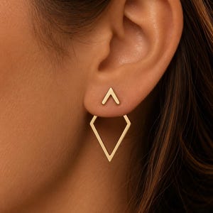 Puede incluir: Un pendiente tipo ear jacket de oro que presenta un pequeño perno en forma de chevrón sobre el lóbulo y un marco abierto en forma de diamante más grande que cuelga debajo, creando un efecto geométrico superpuesto.
