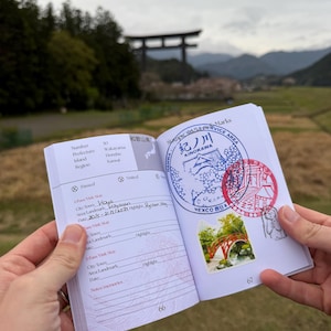 Puede incluir: Un pasaporte abierto muestra sellos y entradas manuscritas. El pasaporte se sostiene frente a un fondo escénico al aire libre con una puerta torii japonesa tradicional y montañas. Los sellos incluyen texto japonés y una imagen de puente.