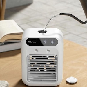 May include: A white portable mini air cooler with a black control panel featuring the text Mini air cooler and two circular icons. The device has a front grille with a visible black fan inside and a top opening where water is being poured in.
