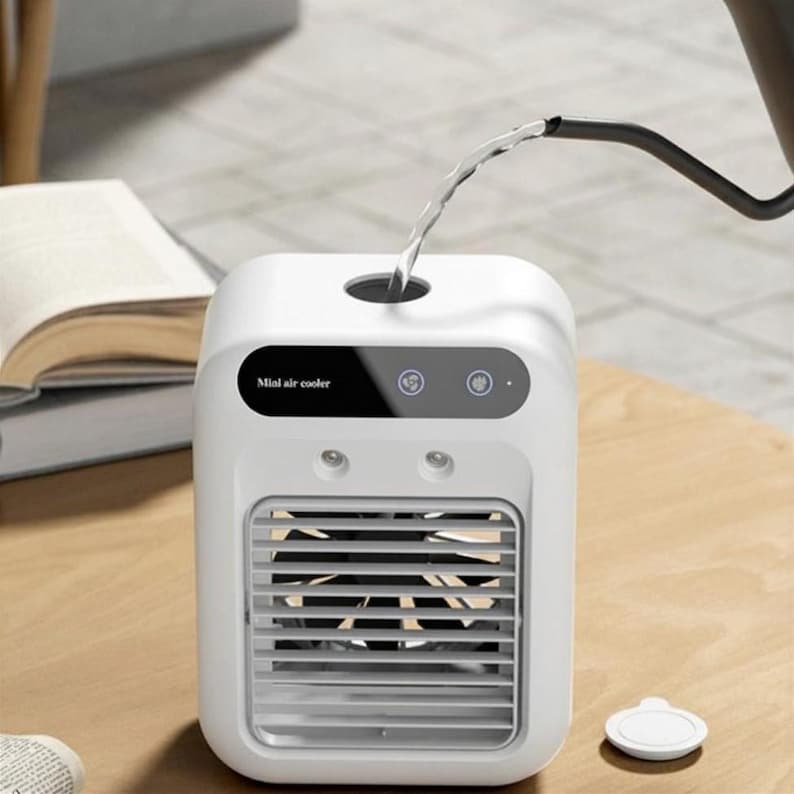 May include: A white portable mini air cooler with a black control panel featuring the text Mini air cooler and two circular icons. The device has a front grille with a visible black fan inside and a top opening where water is being poured in.
