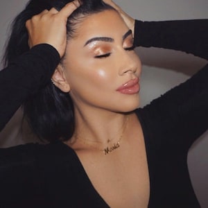 Könnte beinhalten: Nahaufnahme einer Person mit dunklem Haar und einer goldenen Halskette mit dem Namen "Maria". Die Person trägt ein schwarzes Oberteil und hat die Augen geschlossen. Das Make-up umfasst goldenen Lidschatten und rosa Lippenstift.