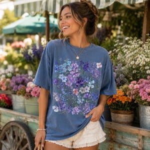 May include: A blue t-shirt featuring a floral design in shades of purple, blue, and white. The shirt is worn by a person standing in front of a flower stand. The person is wearing white shorts and a gold necklace.