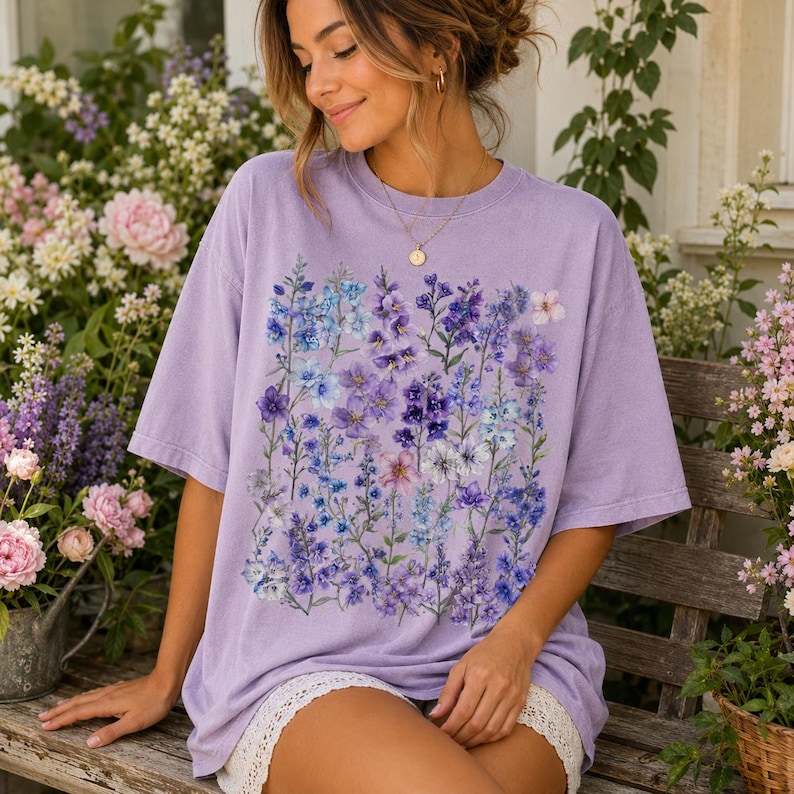 May include: A lilac-colored t-shirt featuring a vibrant floral design in shades of purple, blue, and white. The graphic tee has a relaxed fit and short sleeves. The shirt is worn by a person sitting on a wooden bench.