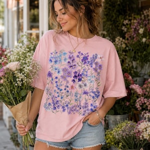 May include: A light pink t-shirt featuring a floral design in shades of purple and blue. The short-sleeved shirt has a crew neck and a relaxed fit. The floral pattern is centered on the front of the shirt. The shirt is worn with denim shorts.