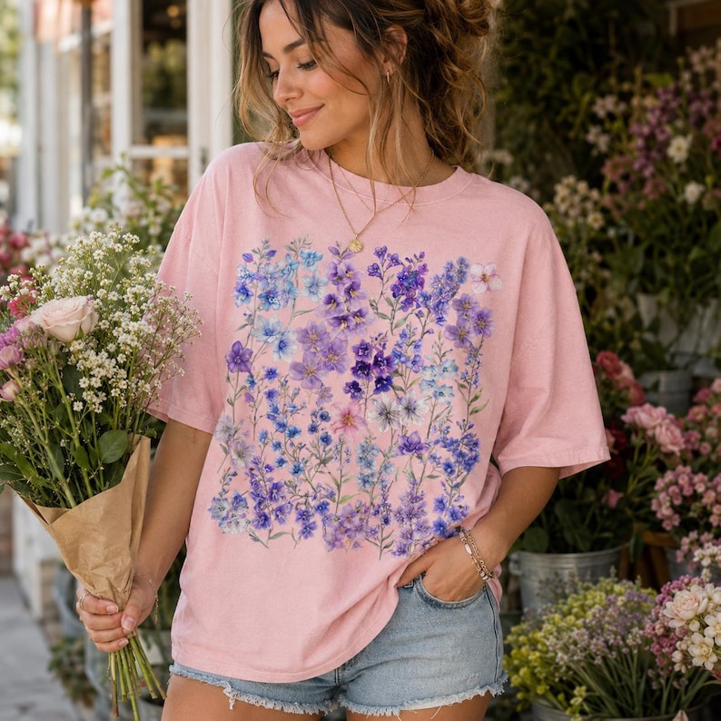 May include: A light pink t-shirt featuring a floral design in shades of purple and blue. The short-sleeved shirt has a crew neck and a relaxed fit. The floral pattern is centered on the front of the shirt. The shirt is worn with denim shorts.