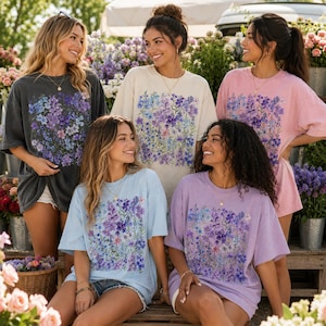 May include: Five women wearing oversized t-shirts with floral designs. The shirts come in various colors: charcoal, cream, light blue, lavender, and pink. The floral print features purple, blue, and white flowers. The women are smiling and posing outdoors.