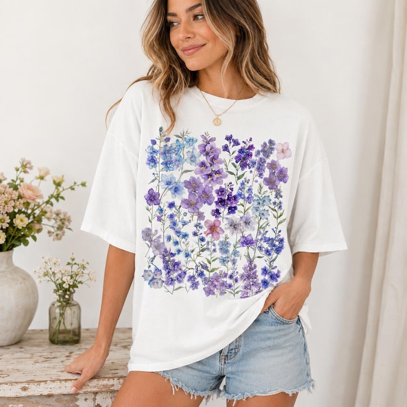May include: White t-shirt featuring a square floral design in shades of purple and blue. The design includes various flower types and green stems. The shirt has short sleeves and a relaxed fit.