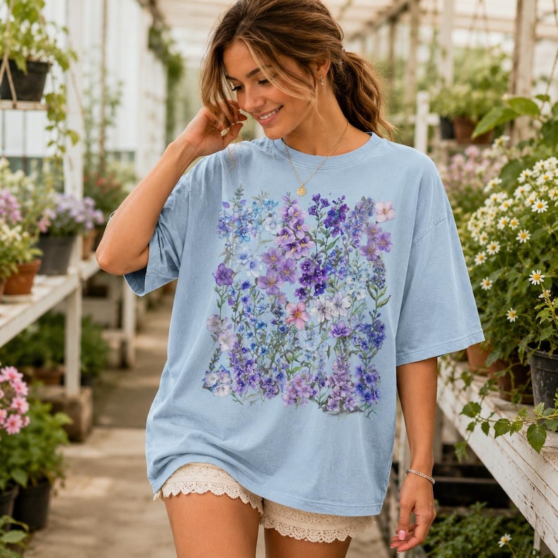 May include: A light blue t-shirt featuring a floral design in shades of purple, blue, and pink. The oversized tee has short sleeves and a crew neck. The floral pattern is centered on the front of the shirt.