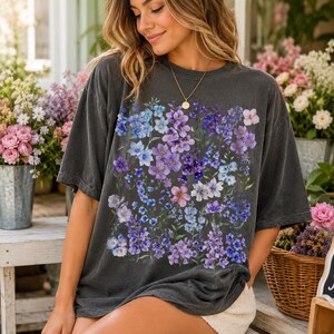 May include: Dark gray t-shirt featuring a vibrant floral design in shades of purple, blue, and pink. The graphic tee has a relaxed fit and short sleeves. The floral pattern is centered on the front of the shirt.