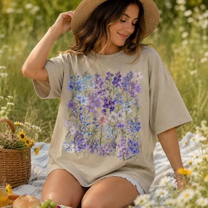 May include: A beige t-shirt featuring a floral design in shades of purple and blue. The shirt is worn by a person sitting on a blanket in a field of flowers. A straw hat and a picnic basket are also visible.