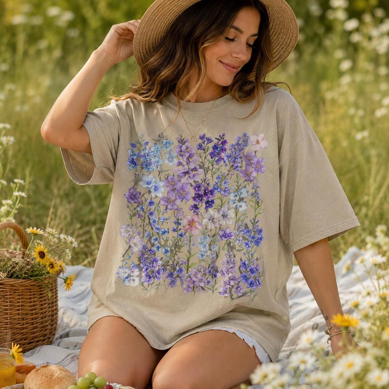 May include: A beige t-shirt featuring a floral design in shades of purple and blue. The shirt is worn by a person sitting on a blanket in a field of flowers. A straw hat and a picnic basket are also visible.
