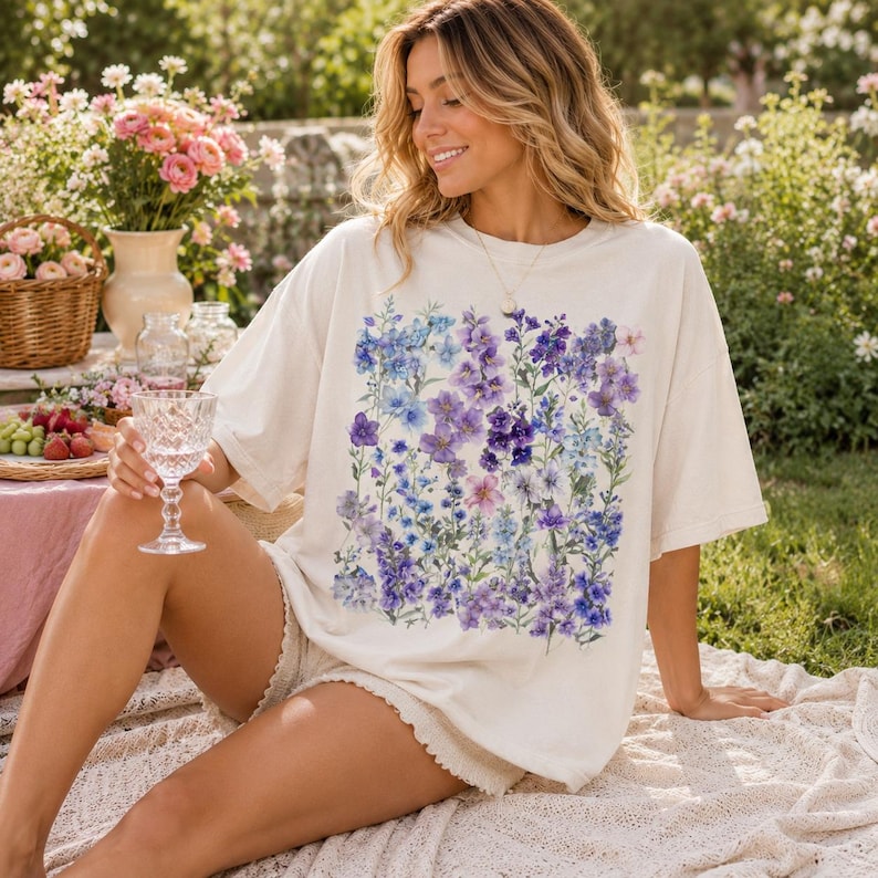 May include: Cream-colored t-shirt featuring a vibrant floral design in shades of blue, purple, and pink. The shirt is worn with white shorts. The image is taken outdoors with a picnic setting in the background.