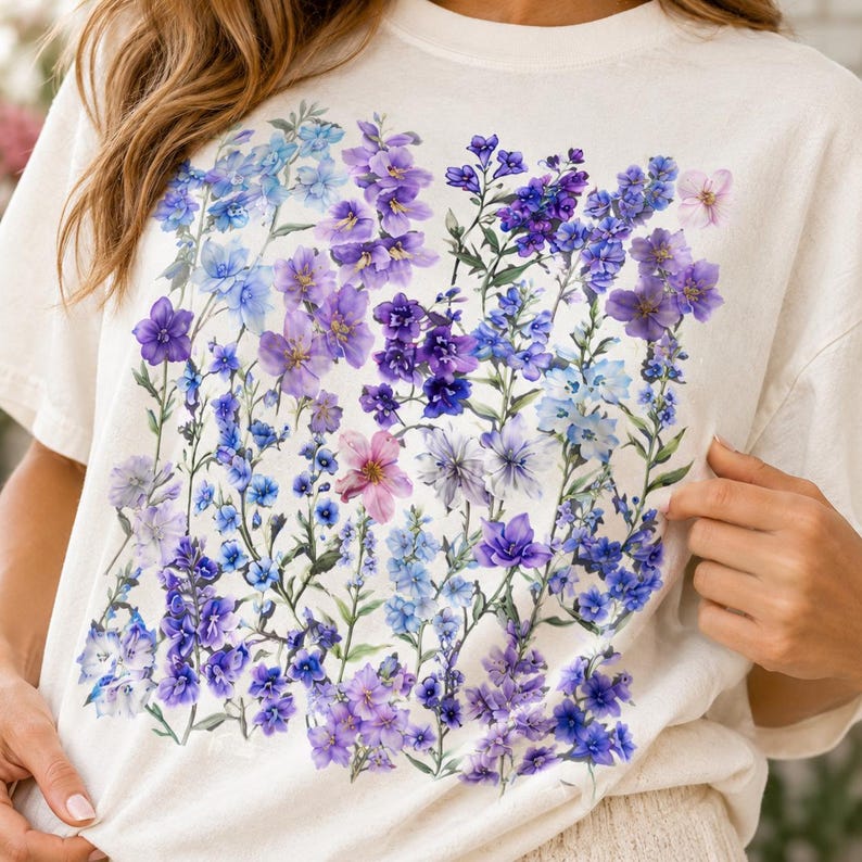 May include: Cream-colored t-shirt featuring a vibrant floral design. The design showcases a variety of purple, blue, and pink flowers with green stems and leaves. The flowers are arranged in a square pattern across the front of the shirt.