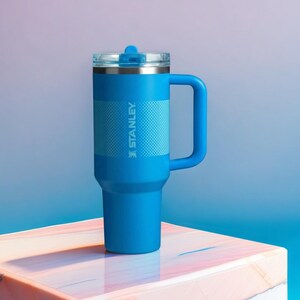 May include: A blue Stanley tumbler with a handle and a clear lid. The tumbler has a textured band around the middle and the Stanley logo printed on the side. The background is a gradient of blue and purple.