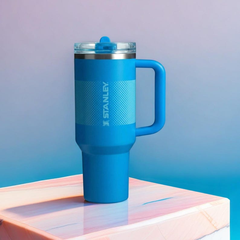 May include: A blue Stanley tumbler with a handle and a clear lid. The tumbler has a textured band around the middle and the Stanley logo printed on the side. The background is a gradient of blue and purple.