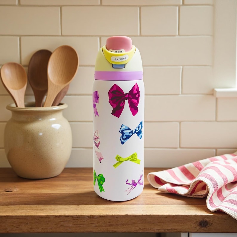 May include: White insulated water bottle decorated with colorful bow illustrations. The bottle has a pink push button lid with the text "Lift to Unlock". The bottle is on a wooden surface with wooden spoons in a ceramic jar.